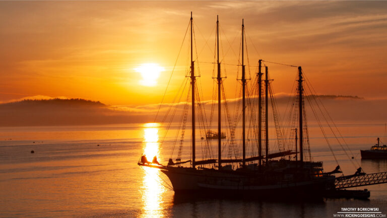 Taken on the morning of July 4th, 2018, this Bar Harbor photo captures a four-masted schooner silhouetted against a brilliant golden sunrise. Low-lying coastal fog rests over the islands in the distance as morning light reflects across the calm water.